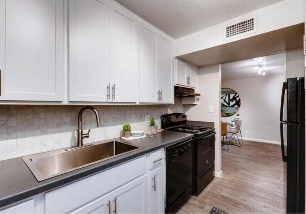 An interior view of a kitchen in Scottsdale apartment for rent at The Glen at Old Town in Scottsdale, AZ featuring kitchen cabinetry, kitchen sink, fridge, stovetop, and oven, and a partial view of a dining room area in the apartment featuring a ceiling fan with a light fixture and a dining table with chairs.