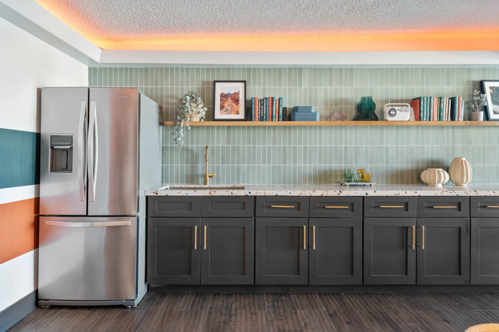 Modern kitchen with a stainless steel refrigerator, dark cabinets, long countertop, open shelf with books and decor, and green tile backsplash. Warm lighting glows from the ceiling above.