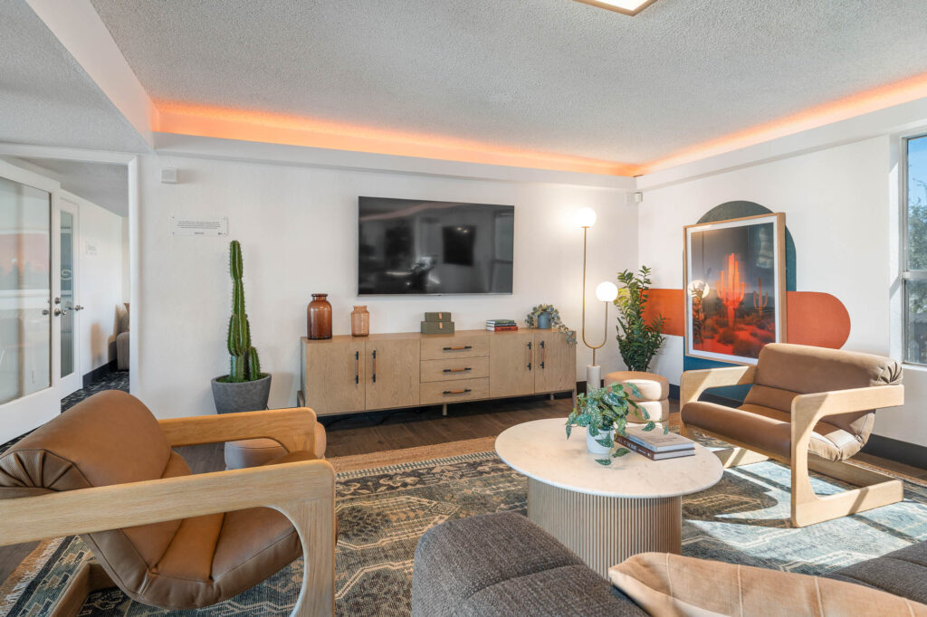 A modern living room with wood-accented furniture, a round coffee table, a large wall-mounted TV, potted plants, framed artwork, and warm ambient lighting along the ceiling.