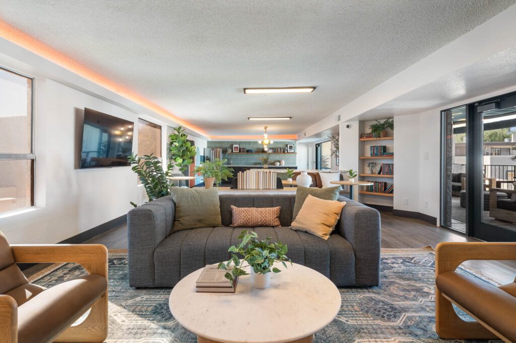 Modern living room with a gray sofa, two brown armchairs, a round coffee table with plants and books, wall-mounted TV, large windows, shelves, and an open kitchen and dining area in the background.