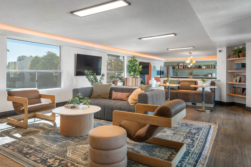 A modern living room with large windows, a gray sofa, light wooden chairs, round coffee table, houseplants, bookshelves, and an open kitchen with barstools in the background. Warm lighting creates a cozy atmosphere.