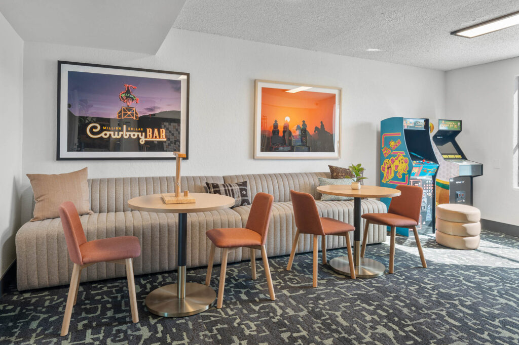 A modern lounge with a striped beige sofa, round wooden tables, and orange chairs. Two framed artworks hang on the wall. In the corner, there are two arcade machines and a stack of round cushions. The room has patterned carpet flooring.