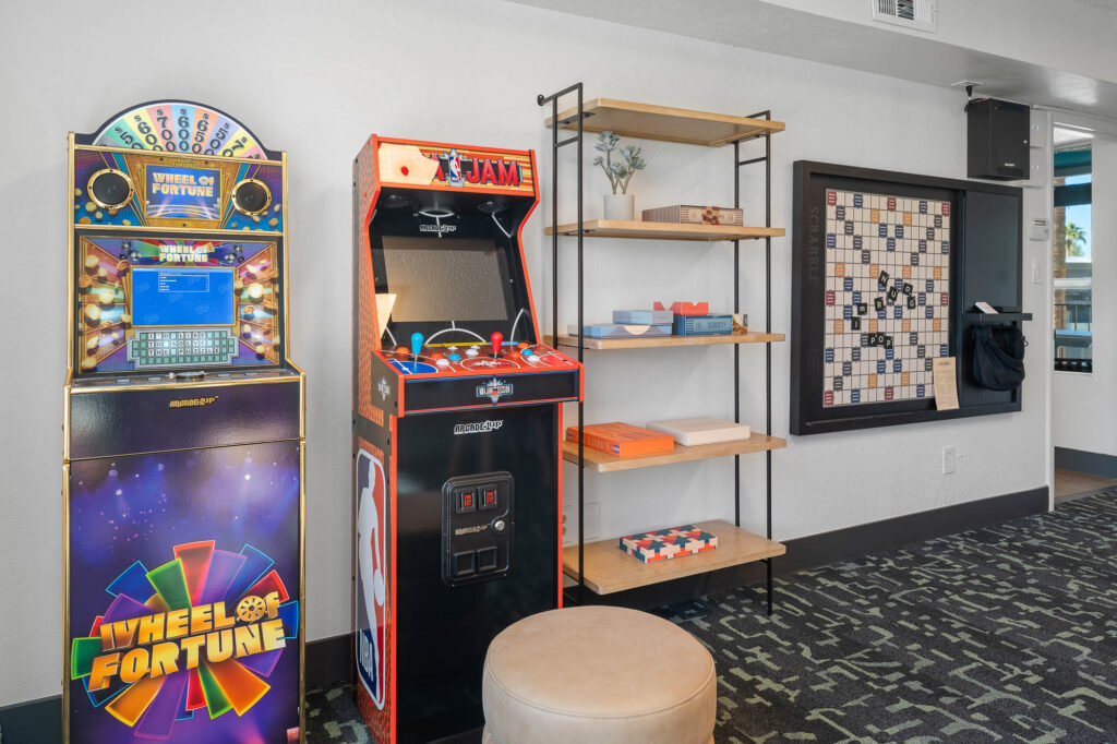 A small game room with a Wheel of Fortune slot machine, an NBA Jam arcade game, a shelving unit with games and décor, a giant wall Scrabble board, and a round beige ottoman on a patterned carpet.