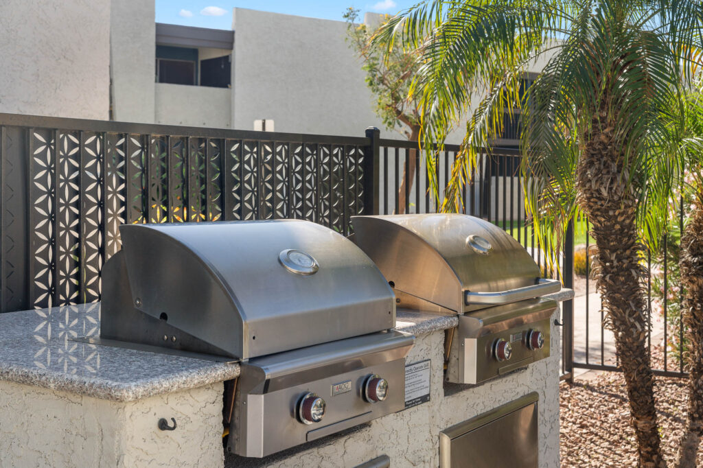 Two stainless steel outdoor grills with closed lids are built into a stone countertop, set beside a black metal fence and a palm tree, with modern white buildings in the background.