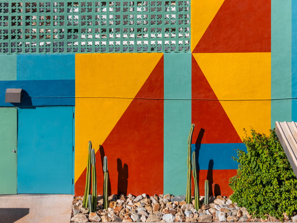 A colorful geometric mural with teal, yellow, red, and green sections covers a wall above a blue door, with tall cacti and rocks planted in front. Bright sunlight casts shadows on the vivid design.