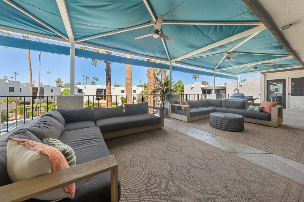 A shaded outdoor lounge area with modern gray sofas, a round ottoman, and ceiling fans overlooks a swimming pool and palm trees, with apartment buildings visible in the background.