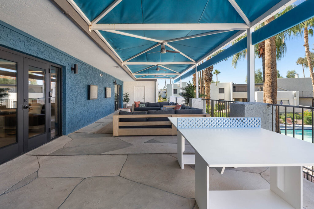 A covered outdoor patio with blue awnings, a white ping pong table, modern couches, and palm trees visible near a swimming pool in the background.