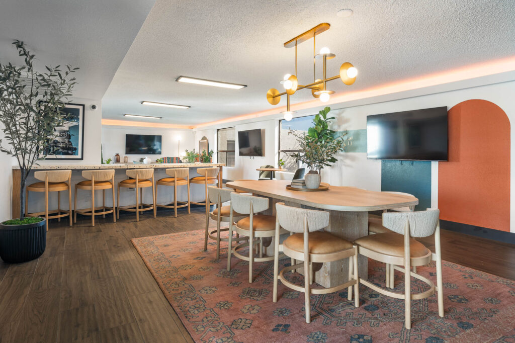 A modern dining area with a wooden table and eight chairs on a patterned rug, two wall-mounted TVs, a bar counter with six stools, and indoor plants. Gold light fixture and colorful wall accents add a stylish touch.