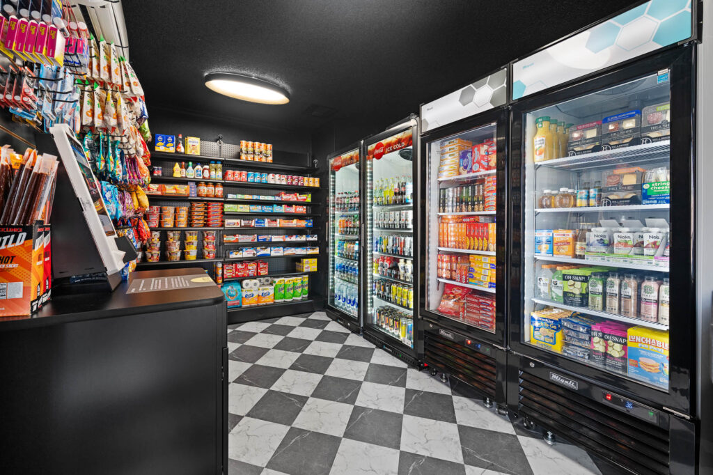 A small convenience store with black-and-white checkered floor, refrigerated cases filled with drinks and snacks, shelves stocked with packaged foods, and a self-checkout kiosk on the left.