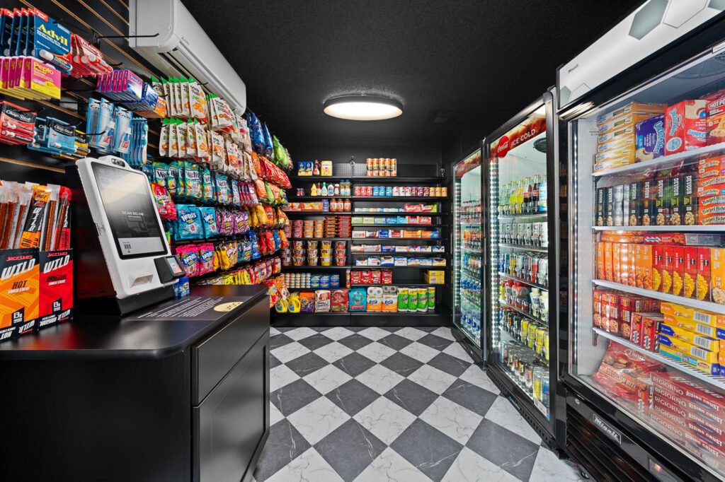 A small convenience store with black-and-white checkered floors, shelves stocked with snacks, a self-checkout kiosk, and refrigerators filled with drinks and packaged food. Bright lighting highlights the colorful products.