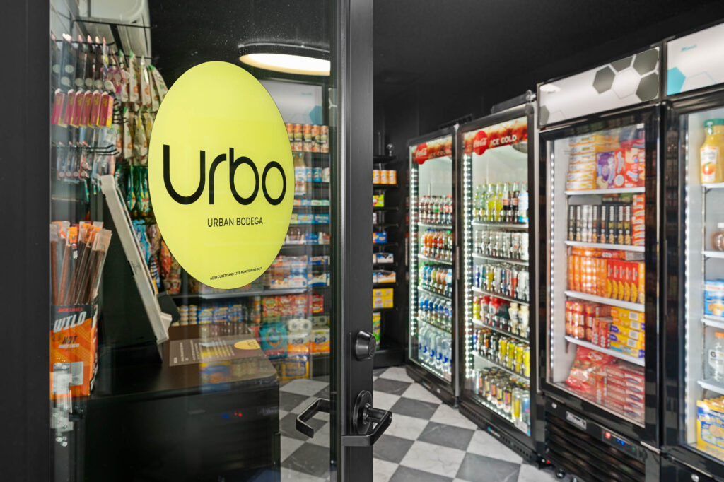 Glass door with a yellow Urbo Urban Bodega sticker opens to a brightly lit convenience store. Inside, shelves and coolers are stocked with drinks, snacks, and groceries on a checkered tile floor.