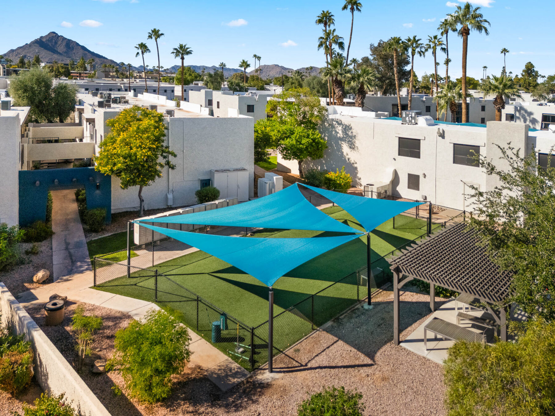 A fenced dog park with artificial grass and large blue shade sails sits among white pet friendly apartments, palm trees, and desert landscaping under a clear blue sky.