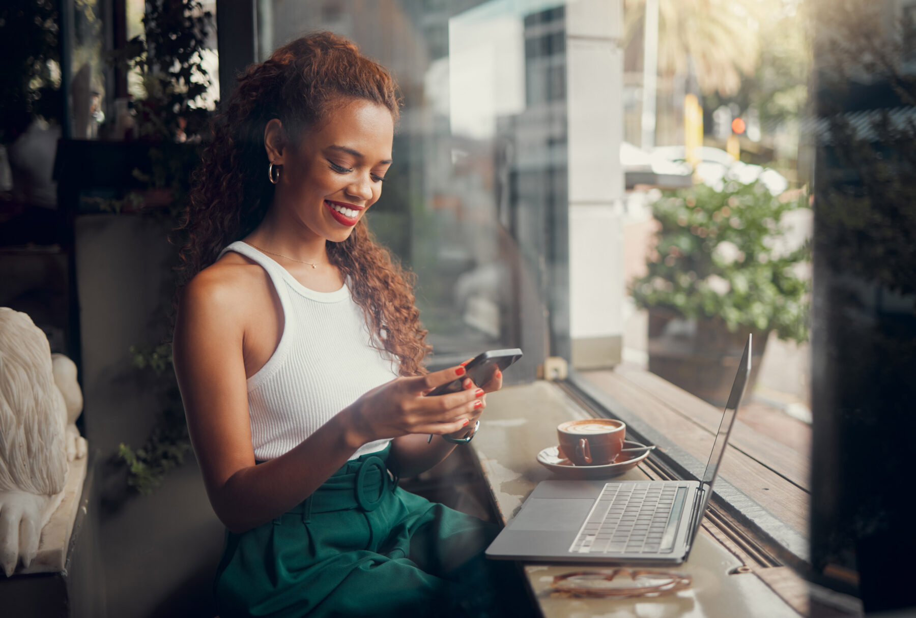 A woman sits by a window in a cafe, smiling at her smartphone. A laptop and a cup of coffee are on the table in front of her. Sunlight streams in, and plants are visible outside the window.